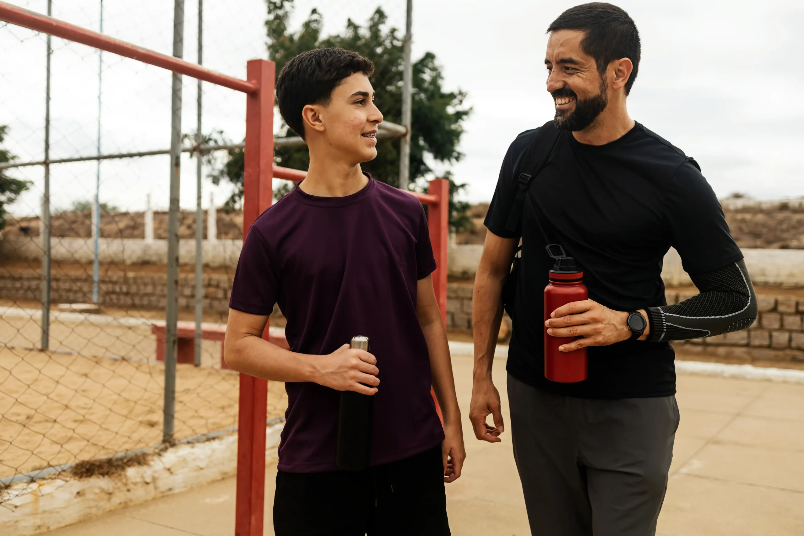 Teenage boy and adult man smiling and talking after outdoor workout with water bottles