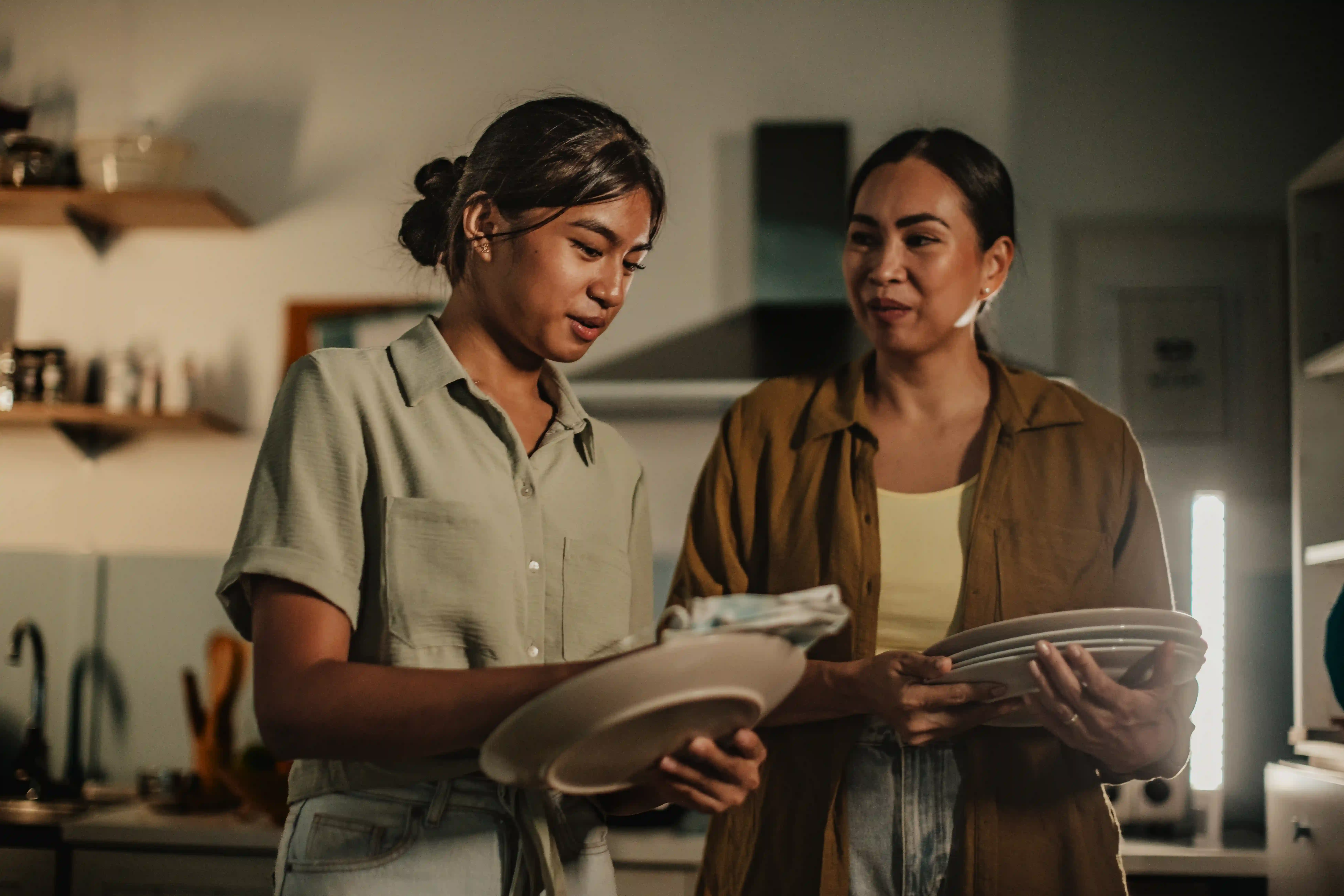 A mother and her teenage daughter share a joyful moment in their cozy kitchen during the evening. They engage in a domestic activity, enjoying each other's company as they prepare.